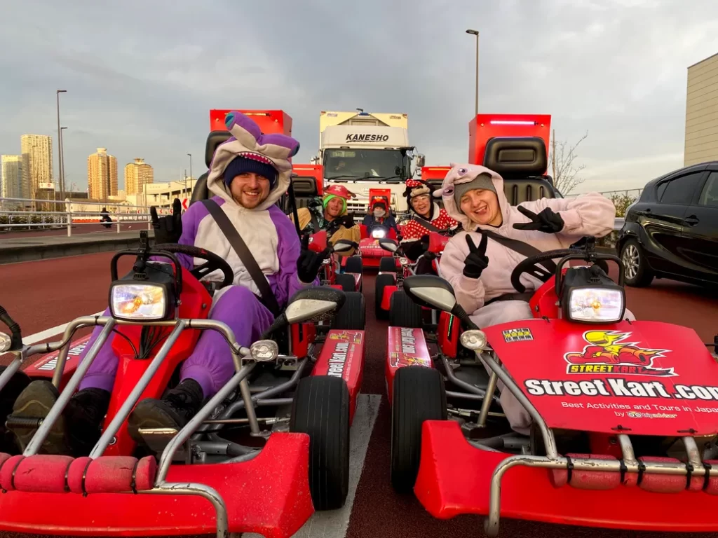People in colorful animal onesies riding red go-karts, posing and waving at the camera in a parking lot with a city skyline in the background.