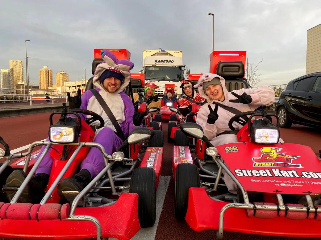 People in colorful animal onesies riding red go-karts, posing and waving at the camera in a parking lot with a city skyline in the background.