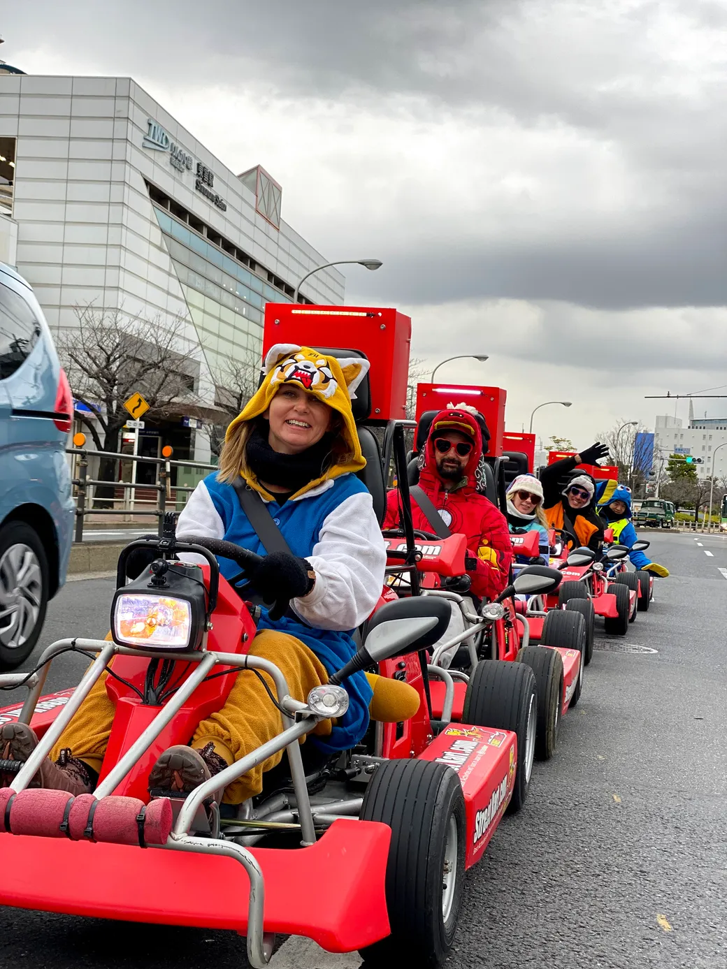 Front rider in a tiger hoodie drives a red pedal go-kart in a parade line on a city street, other costumed riders follow behind.