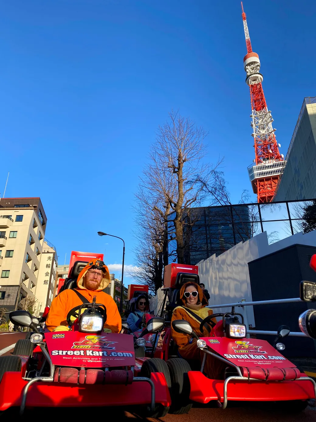 Two people in orange outfits ride red go-karts on a city street, with Tokyo Tower visible in the background.